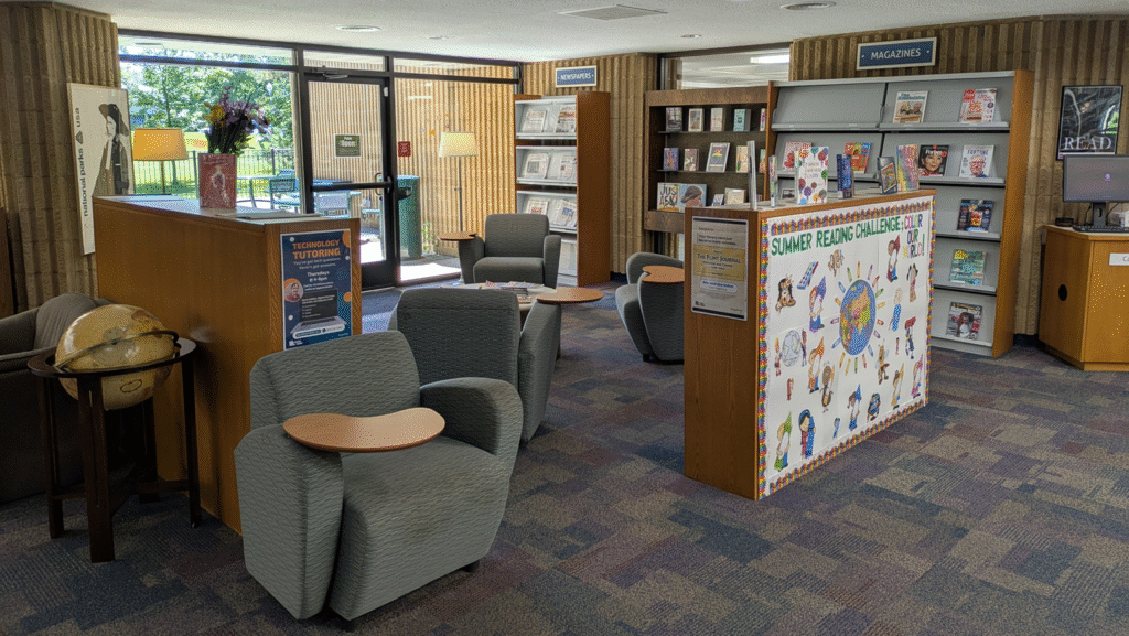 A reading area with chairs with desks attached, newspapers, magazines and a book display.